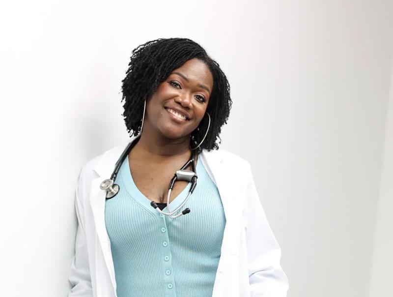 Onye Nnorom leans against a wall, smiling, wearing lab coat and stethoscope.