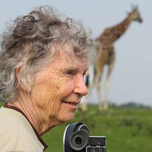 Anne Innis Dagg looks over her camera with a giraffe in the background.