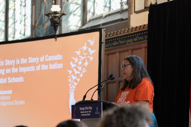 Brenda Wastasecoot speaking at a podium while wearing an orange Every Child Matters shirt.