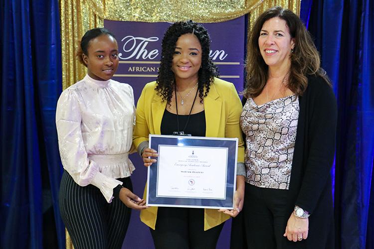 Mariam Olafuyi holds a framed certificate and smiles, flanked by Iris Hategekimana and Kelly Hannah-Moffat