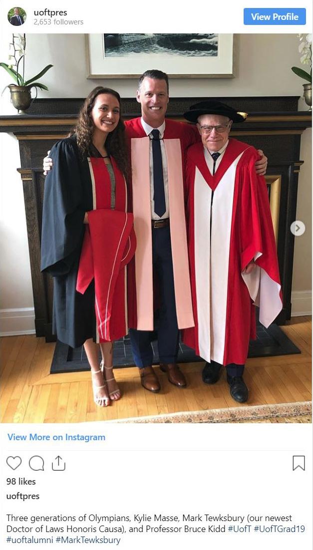 Mark Tewksbury smiles with his arms around Kylie Masse and Bruce Kidd, in front of a fireplace. All three wear academic robes.