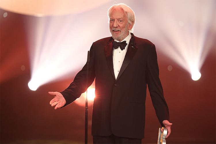 Donald Sutherland smiles and gestures, standing on a stage in spotlights while holding an award.