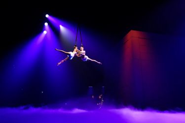 Acrobats in the air above a stage with mist.