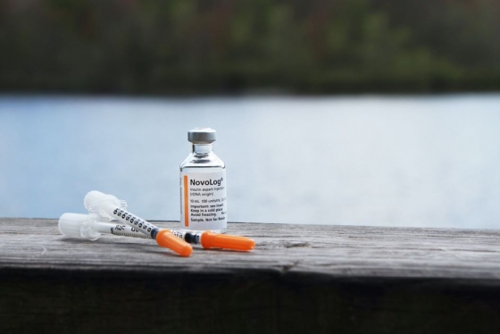 Vial of insulin sits with 2 needles on a wooden table