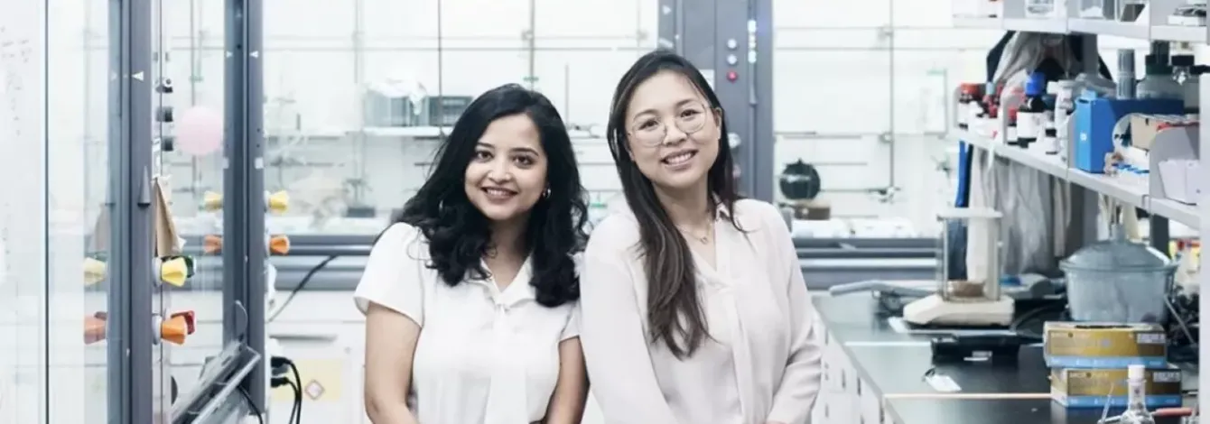 Scientists Nabanita Nawar and Pimyupa Manaswiyoungkul, both wearing white blouses and black pants, stand together in their lab smiling.