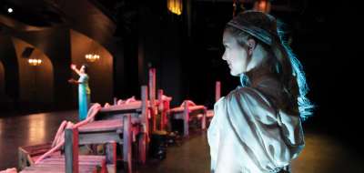 A woman in costume watches from the back of the Hart House Theatre stage as a woman in the spotlight holds up her arms.