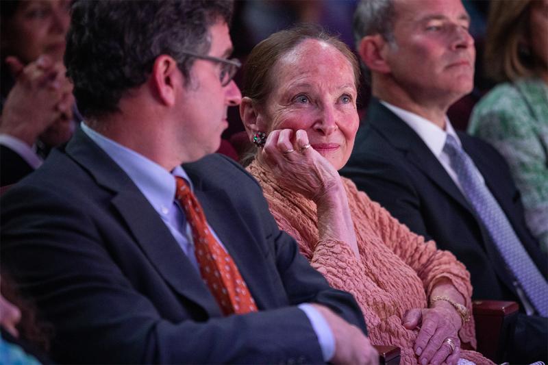 Rosie Abella sitting in an audience, listening, smiling, and resting her chin on one hand.