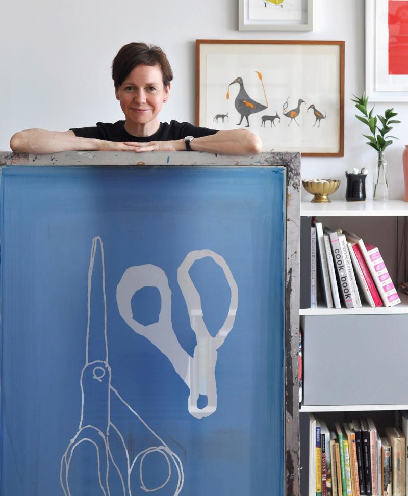 Alanna Cavanagh standing behind a desk in front of her artwork. 