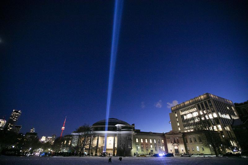 A spotlight shines straight up into the night sky on a snow-covered Front Campus, in front of a lit-up Convocation Hall.