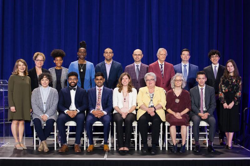 17 winners of Awards of Excellence pose on a stage, standing in two rows for a group picture and smiling. (photo by Gustavo Toledo Photography)