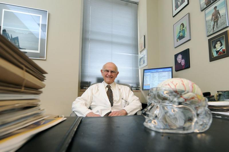 Charles Tator grins as he sits at his office desk, which holds a plastic model of the human brain.