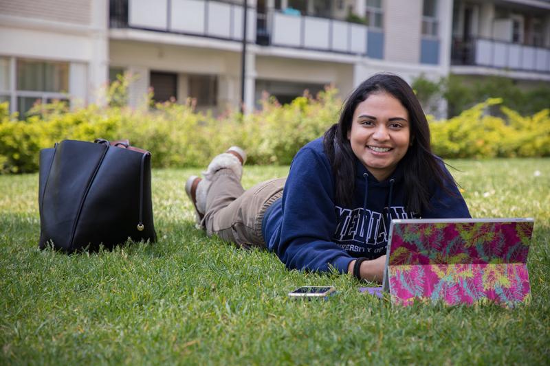 Amanda Khan smiles as she looks up from her laptop while lying on her stomach on a grass lawn.