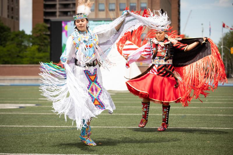 Alanna Pasche and Deanne Hupfield, wearing Indigenous regalia with fringe and embroidery, dance on the turf at Varsity Stadium.