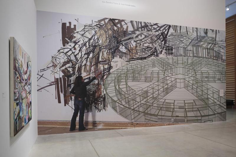 Large painting on wall in gallery showing a woman in jeans and a black shirt with a paint brush painting black, abstract lines. The painting includes a large, circular artistic element with spokes moving in toward the centre, resembling a drawing of the inside of a building.