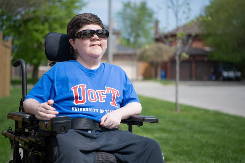 Alex Harold smiles and looks up as he sits outdoors with one hand on his wheelchair controls.