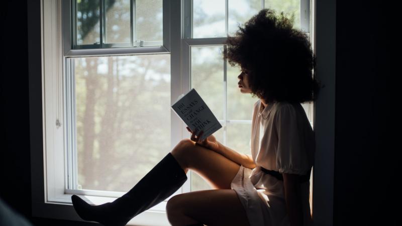 A young woman sits on a sunny windowsill, reading a book.