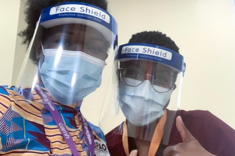 Onye Nnorom and Duate Adegbite, wearing masks and face shields, lean together and give the thumbs up.