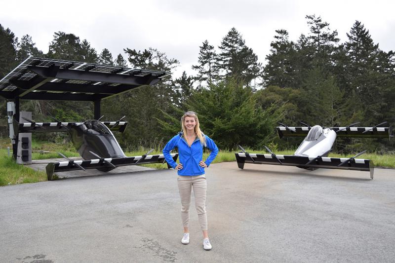 Kristina Menton stands in front of two vehicles that look like race cars with no wheels and very long, wide, flat axles.