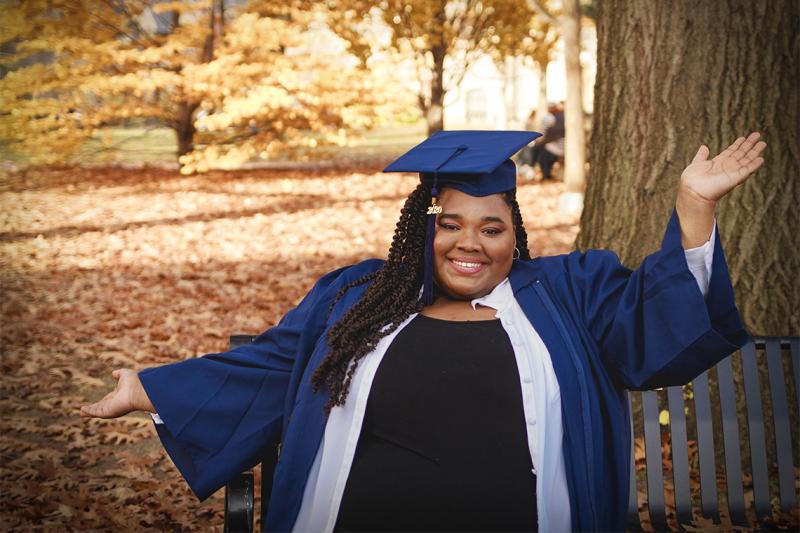 Carisse Samuel stretches out her arms and smiles, as she poses in academic cap and gown on a park bench.
