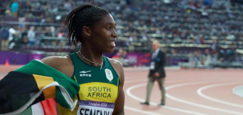 Caster Semenya smiles and waves the South African flag while standing on the track in a stadium.