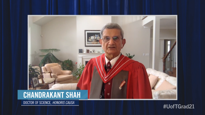 Chandrakant Shah, wearing academic robes, speaks on video from a living room with large plants.