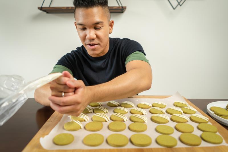 Colin Asuncion smiles as he squeezes icing out of a pastry bag and onto a tray of cookies.