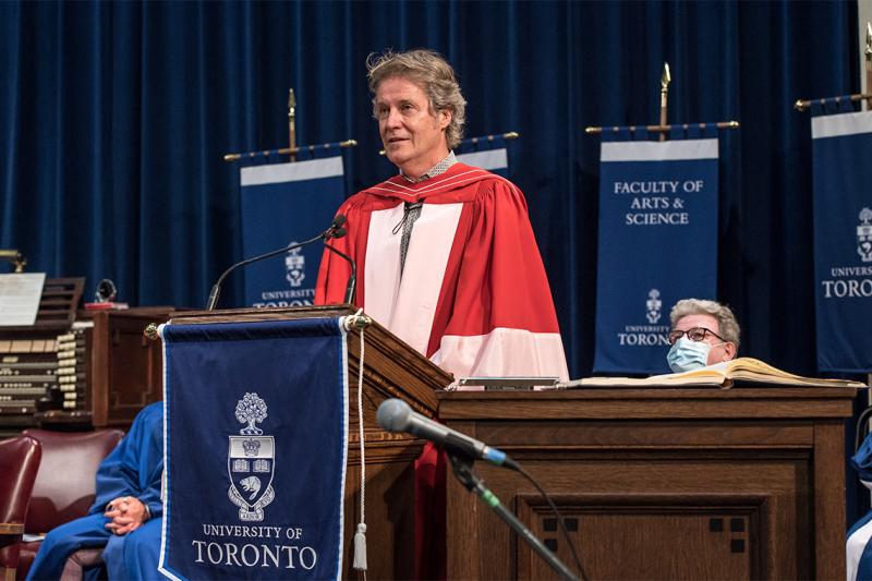 Jim Cuddy, wearing academic robes, speaks at the podium in Convocation Hall.