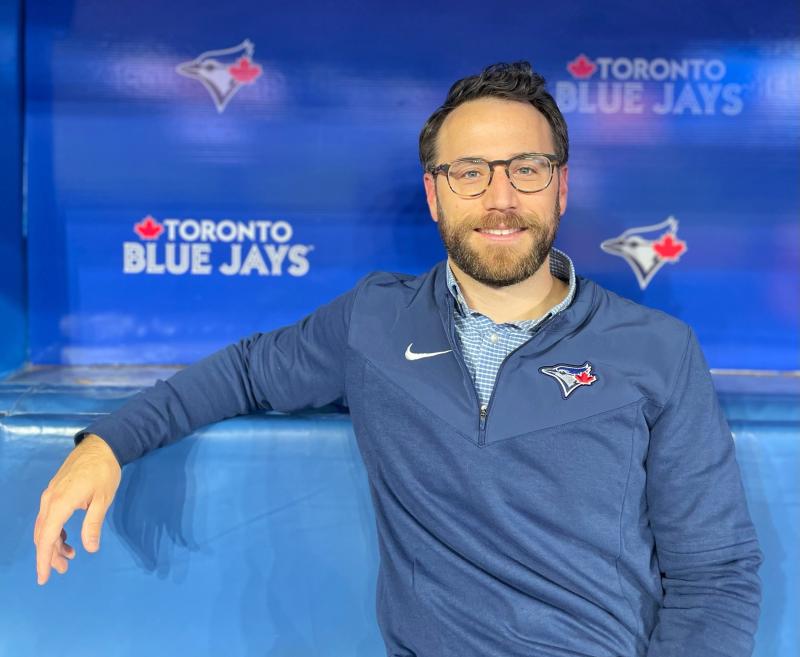 David Lawrence smiles, sitting in the Blue Jays dugout with the team logo behind him.