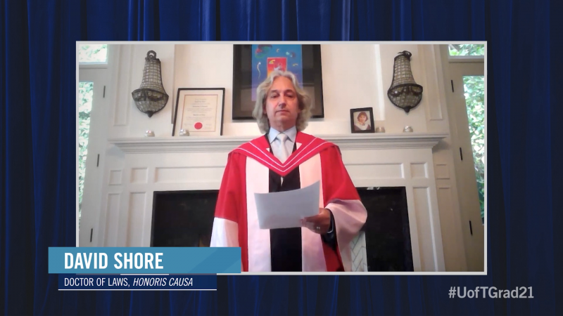 David Shore wears academic robes and speaks in front of a fireplace. On the mantel is his graduation picture as a young man.