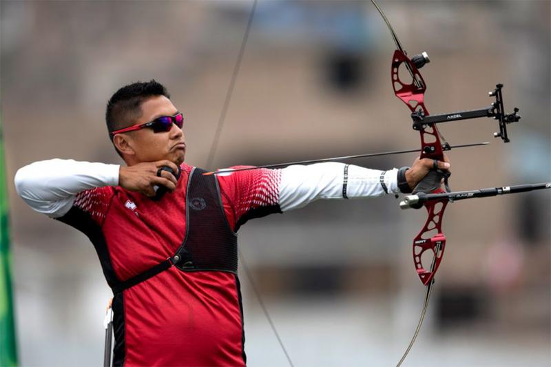 Crispin Duenas in the middle of loosing an arrow from his high-tech archery bow. He wears sunglasses and a chest protector.