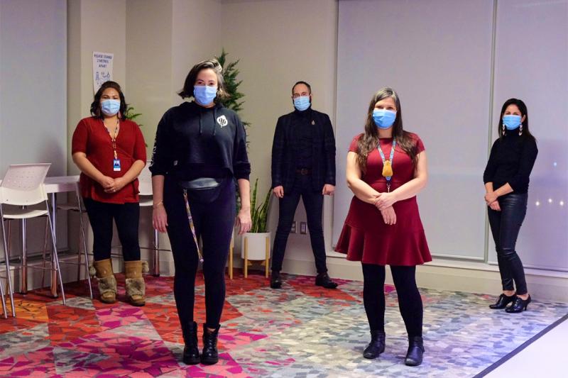 Five people wearing masks, and standing 2 metres apart, pose for a picture on a cosy, colourful carpet.