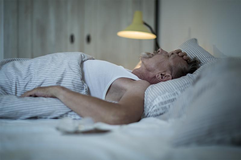A man lies on his back in bed with his eyes closed and one hand on his head.