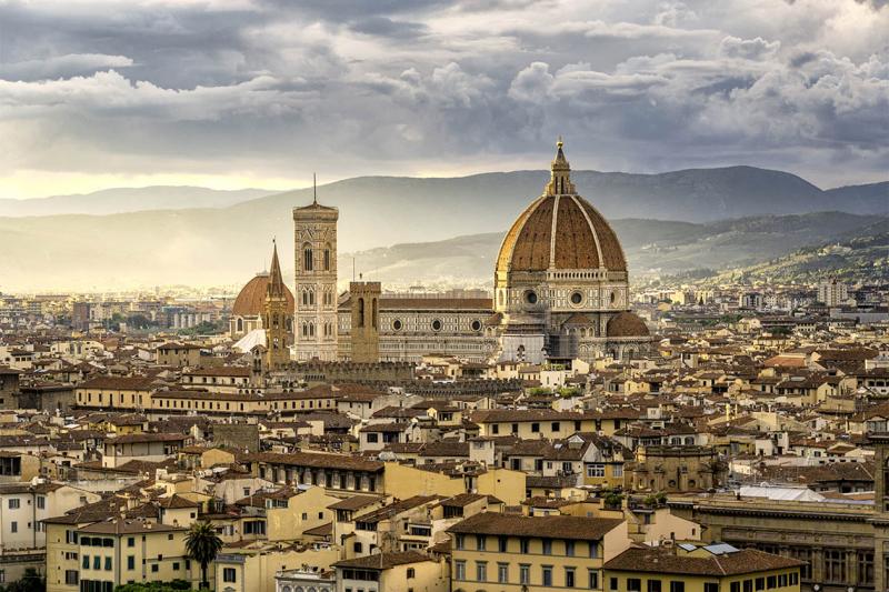 A medieval cathedral decorated in a tile pattern soars above the tiled rooftops of Florence at sunset.