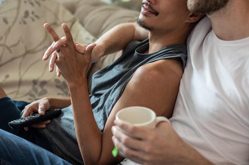 Two men hold hands as one leans comfortably against the other.