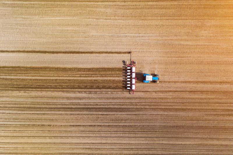Seen from high above, a tractor drags a machine across a crop field, leaving stripes.