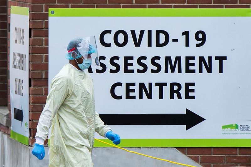 A person in full protective equipment walks by a sign that reads COVID-19 Assessment Centre.