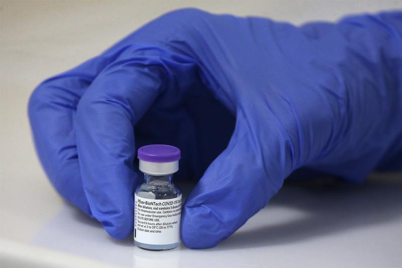 A hand in a medical glove holds a tiny vial. The vial is labelled: Pfizer-BioNTech COVID-19 vaccine.