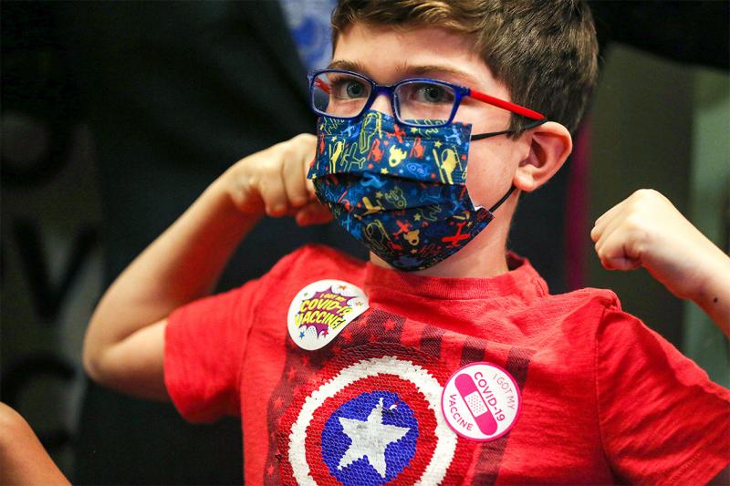 A young boy flexes his muscles. He's wearing a mask, a superhero t-shirt, and a sticker that says I got my COVID-19 vaccine.