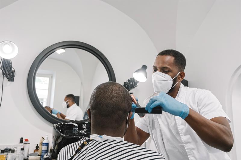 A Black barber in mask and gloves puts the finishing touches on a haircut for a man sitting in a barber chair.