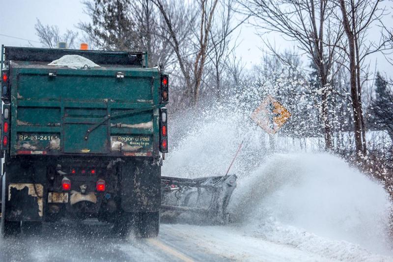 A salt truck with plow sends snow flying over a snowbank at the side of a tree-lined rural road.