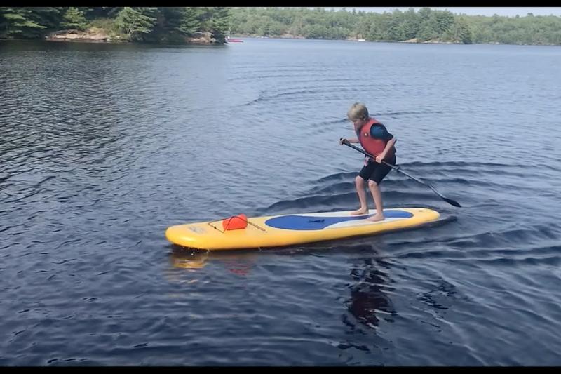 On a lake, a boy stands and jumps on the stern of a paddleboard. Ripples behind show he's moving forward.
