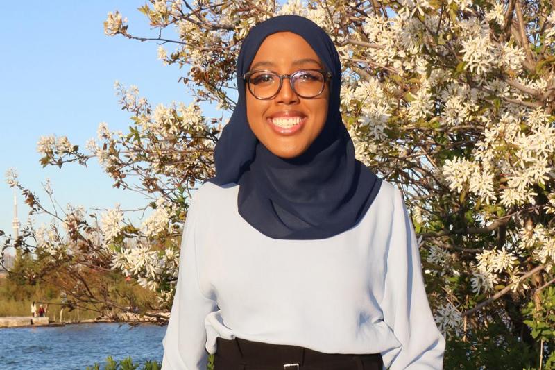 Habon Ali smiles, standing outdoors by a small lake in the woods.