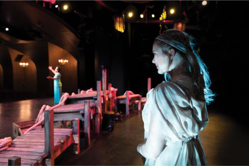 A woman in costume watches from the back of the Hart House Theatre stage as a woman in the spotlight holds up her arms.