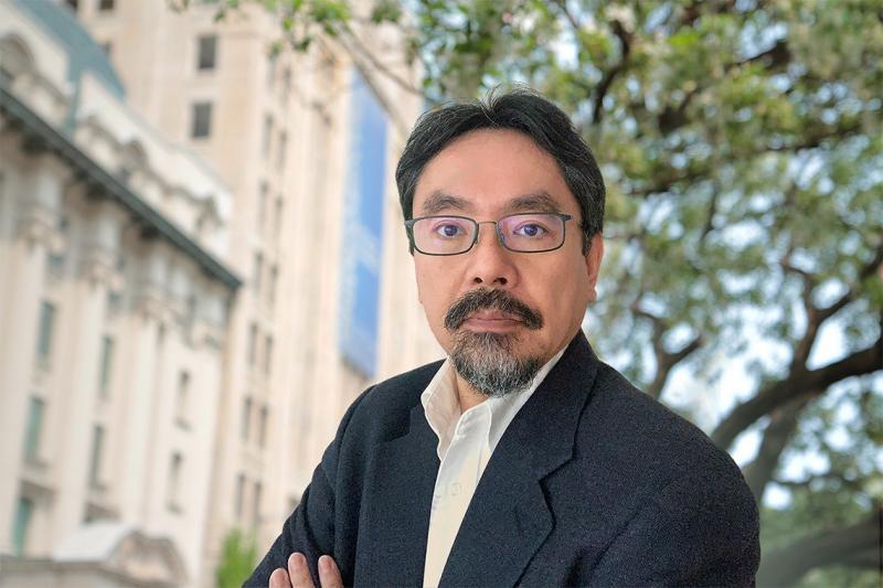 Naoto Hirano looks serious as he stands under a tree outdoors.