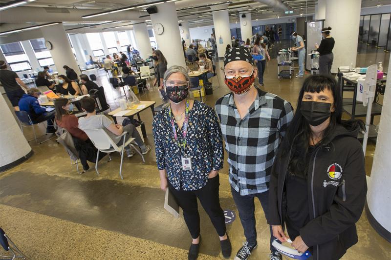 Janet Smylie, Michael Anderson and Suzanne Stewart, wearing masks, stand in an atrium dotted with vaccination stations.