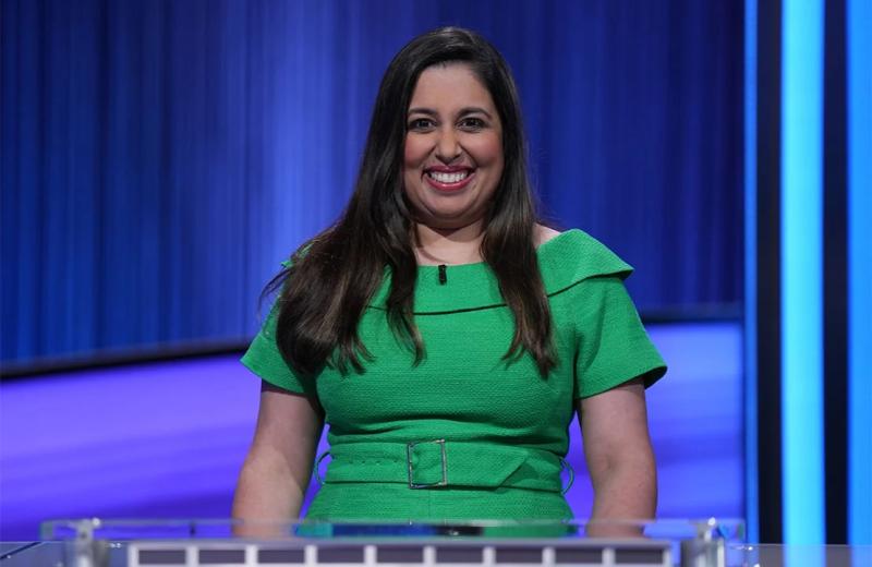 Javeria Zaheer wearing a green dress and smiling on the Jeopardy! set.