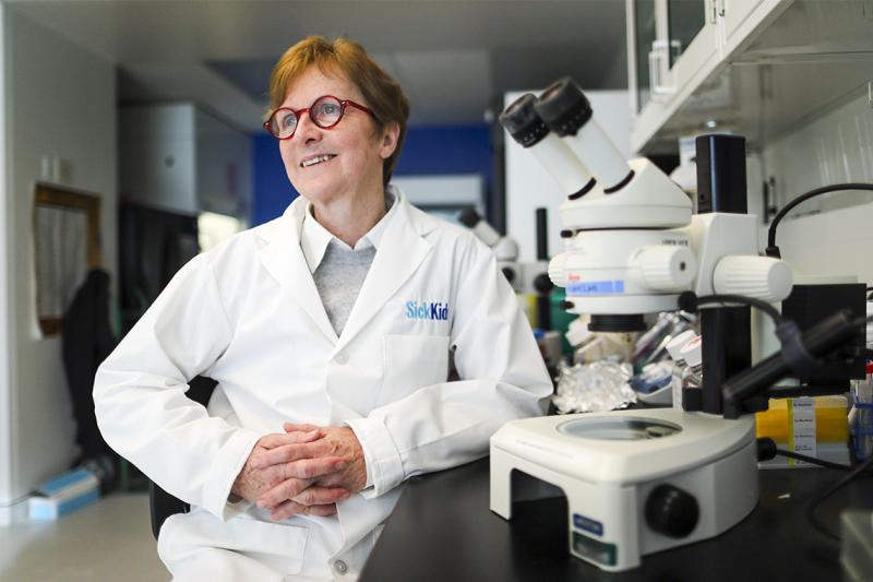 Janet Rossant smiles, wearing a Sick Kids white medical coat and sitting next to a microscope on a bench.