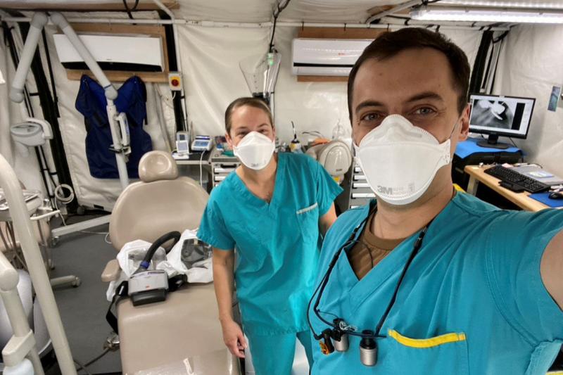 Sgt. Angela Brownell and Jesse Barker, wearing scrubs and masks, stand beside a dental chair inside a large tent.