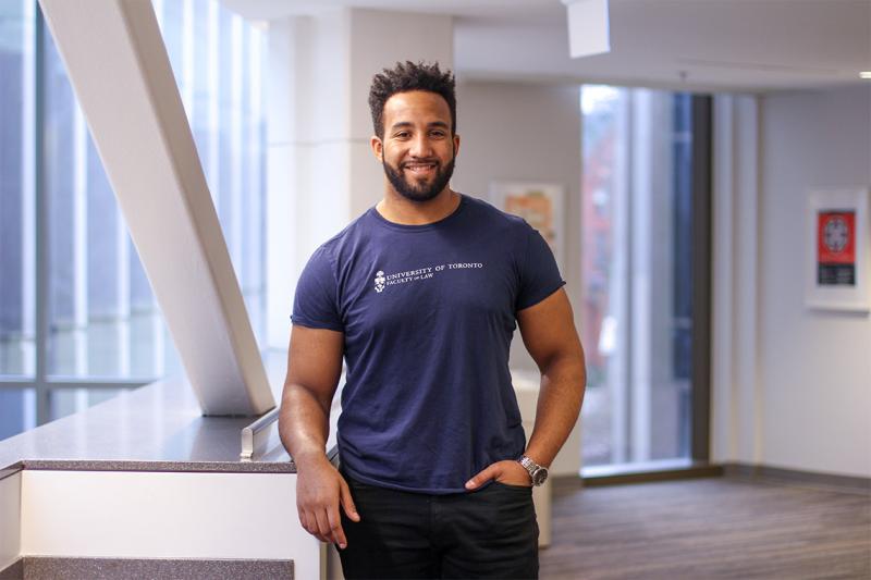 Joshua Lokko smiles, wearing a U of T Faculty of Law T-shirt and standing by an indoor balcony railing.