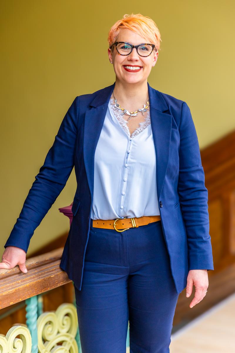 A woman wearing a blue suit standing in front of a bright yellow wall.